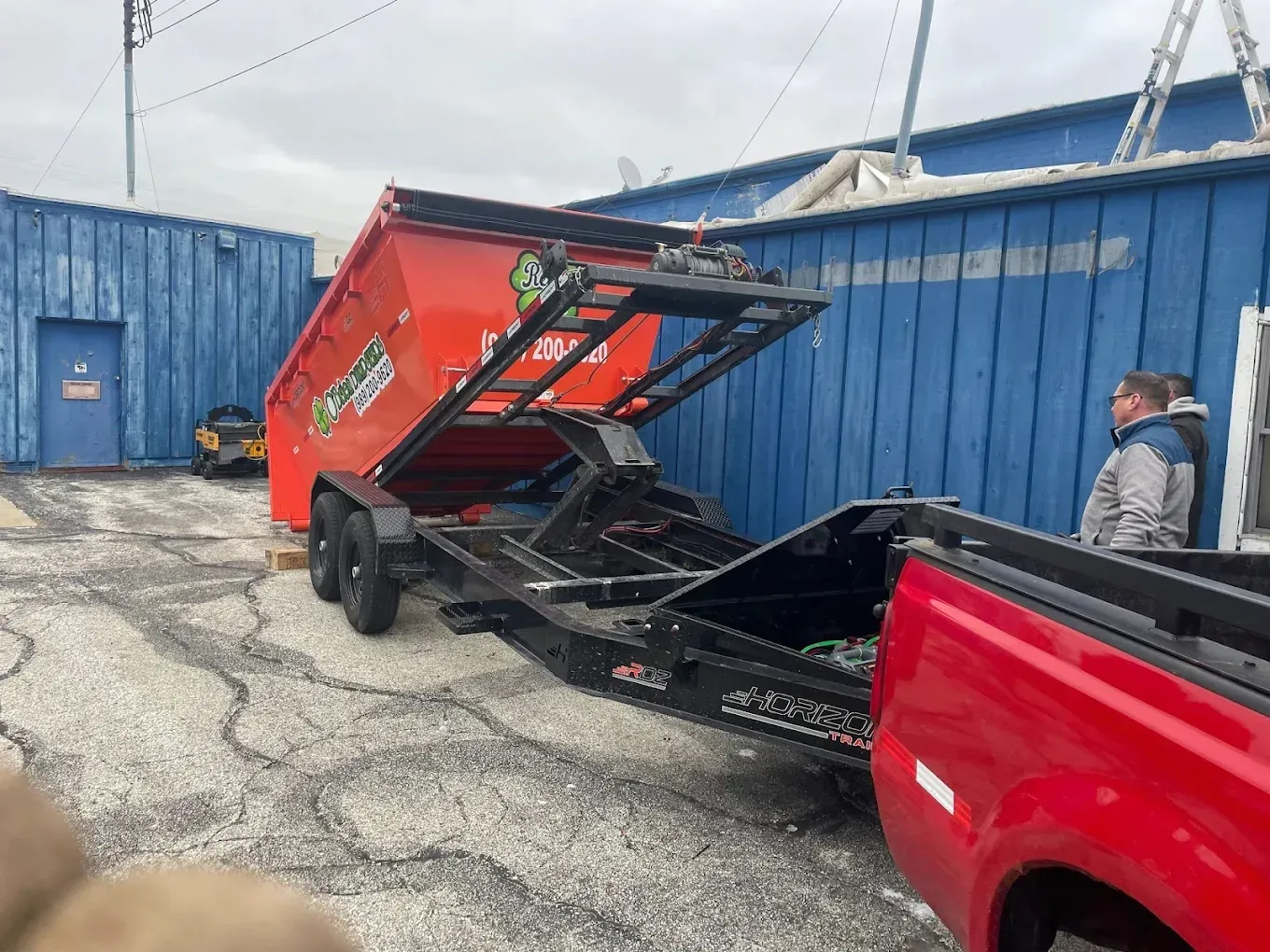 A red dumpster on a trailer being loaded; a red truck is hooked up. Blue buildings are in the background.