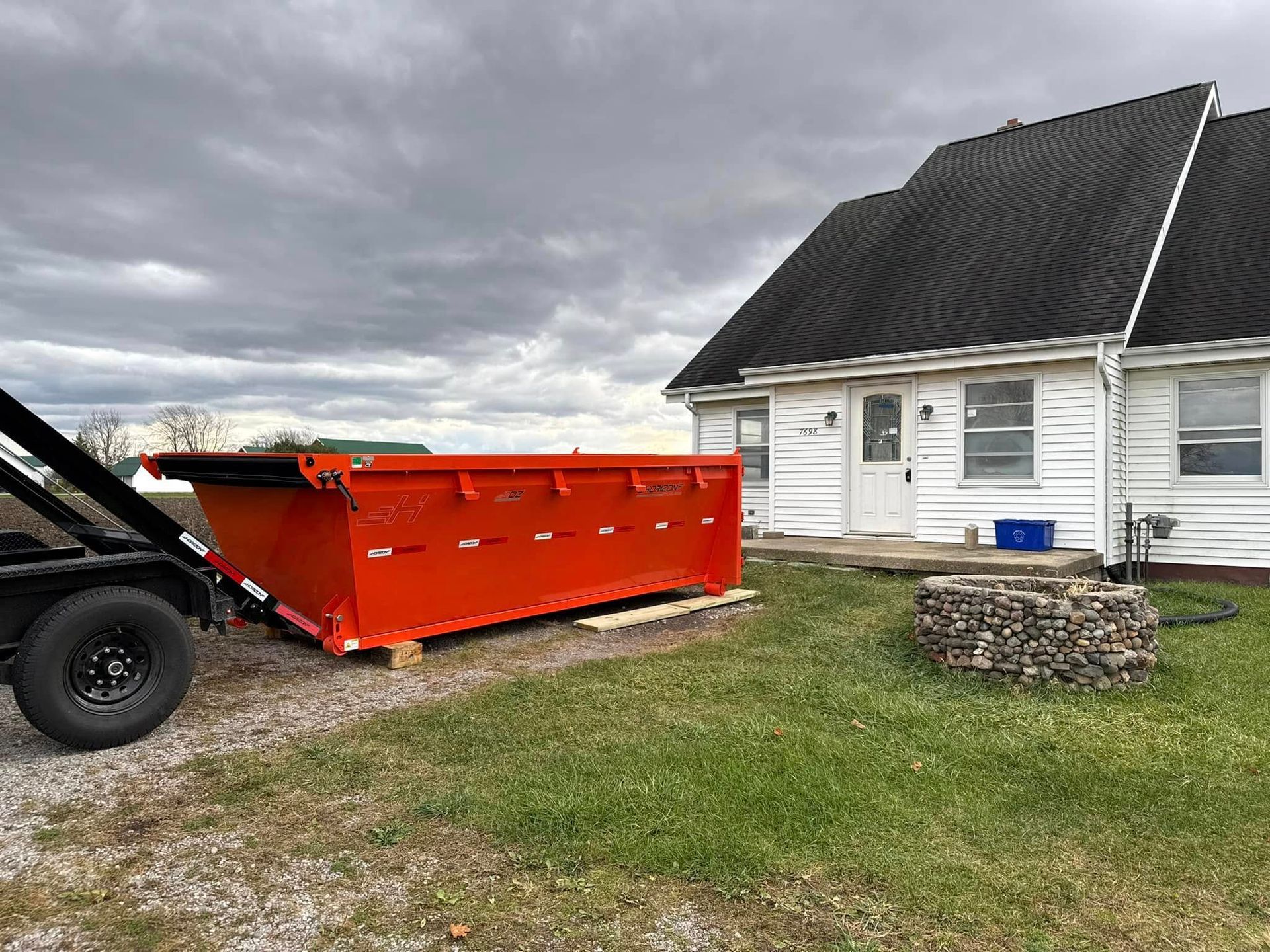 An orange dumpster sits on a trailer next to a white house with a dark roof on a cloudy day.