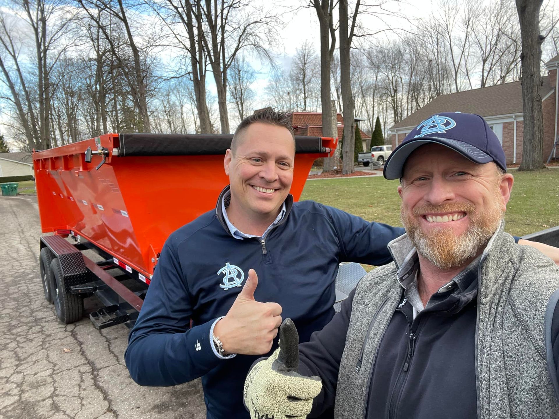 Two men smiling next to an orange dump trailer outdoors. One gives a thumbs up.