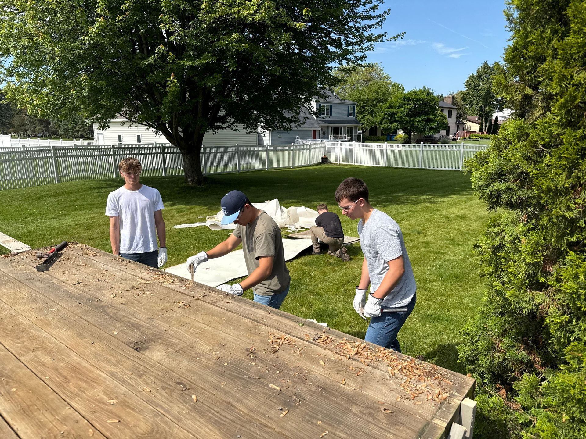 Three people working on a wooden structure outdoors. One wears a cap and uses a tool. Green grass, white fence, and trees in background.