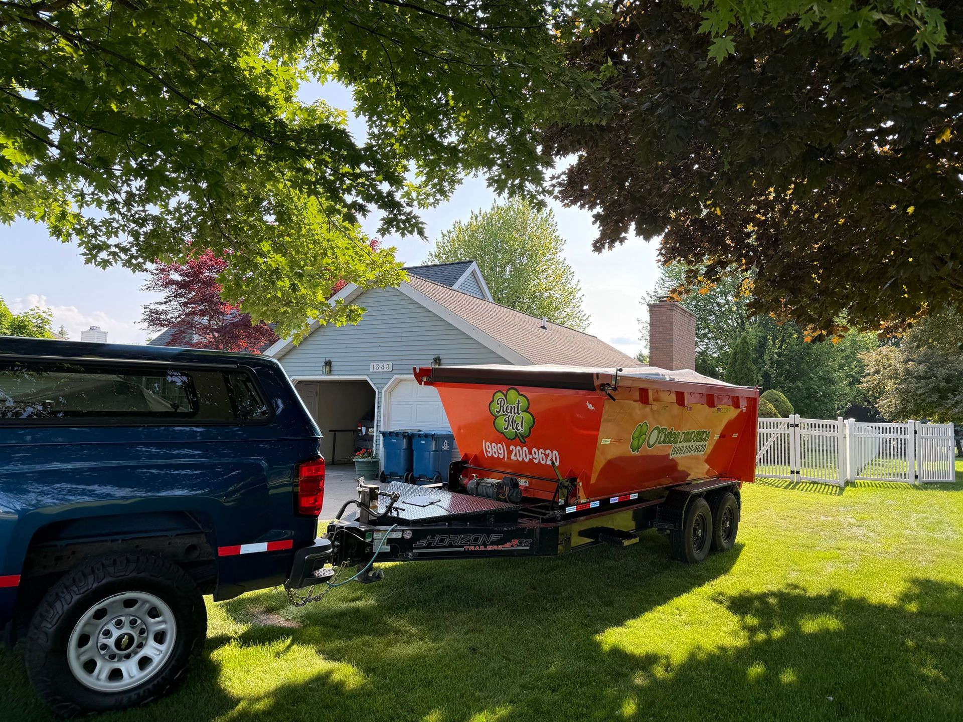 Blue truck towing an orange and yellow trailer with a logo in a grassy yard near a house.