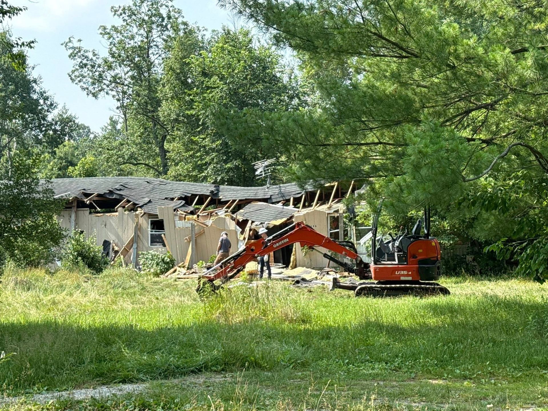 Mini excavator demolishing a house; two people observe. Green trees and grass surround the scene on a sunny day.