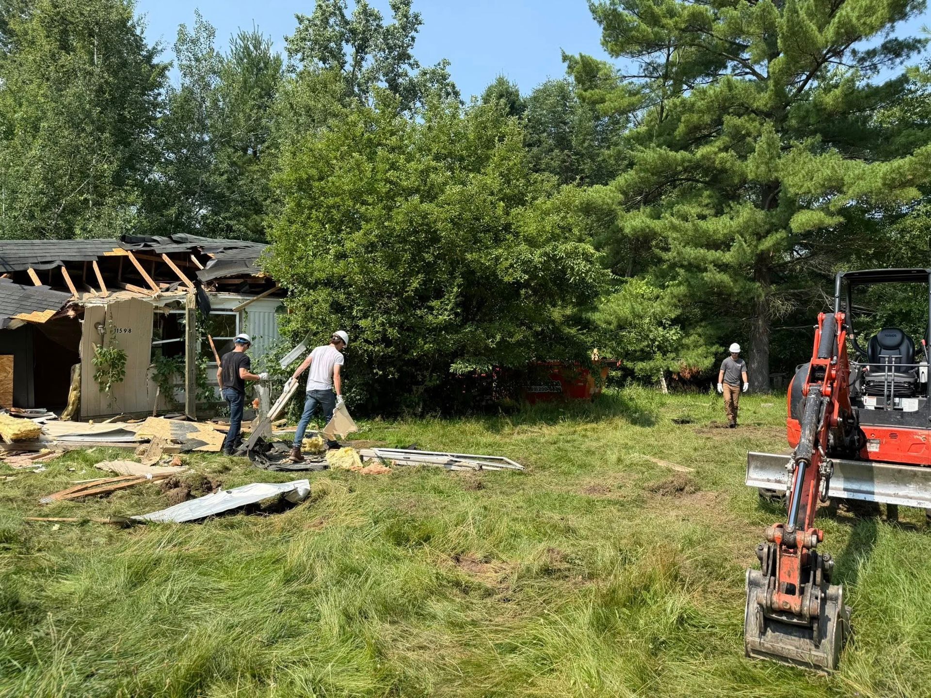 Demolition of a house; workers and excavator on a grassy lot, trees in the background.