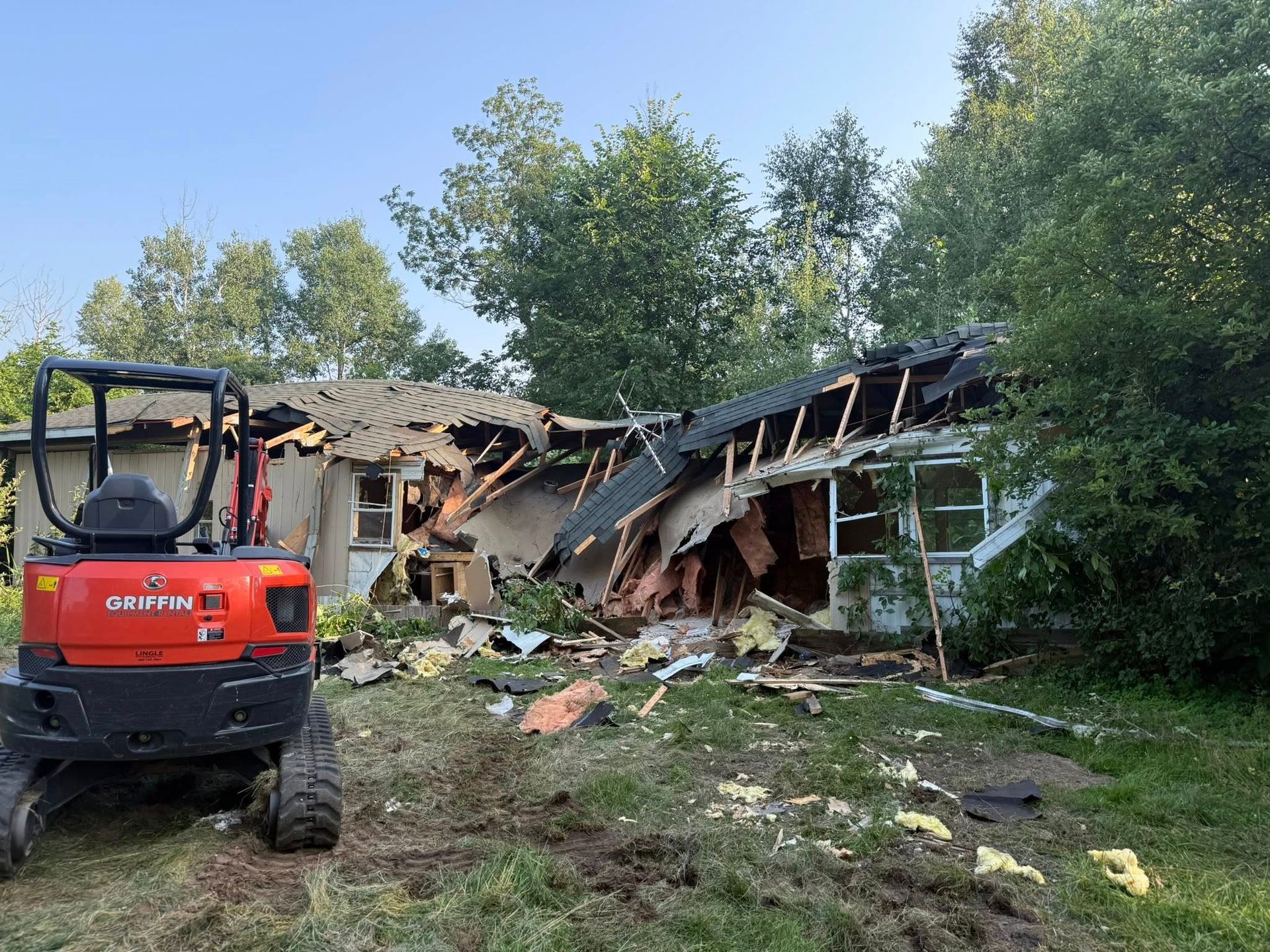 A small excavator sits in front of a partially demolished house with a damaged roof and debris.