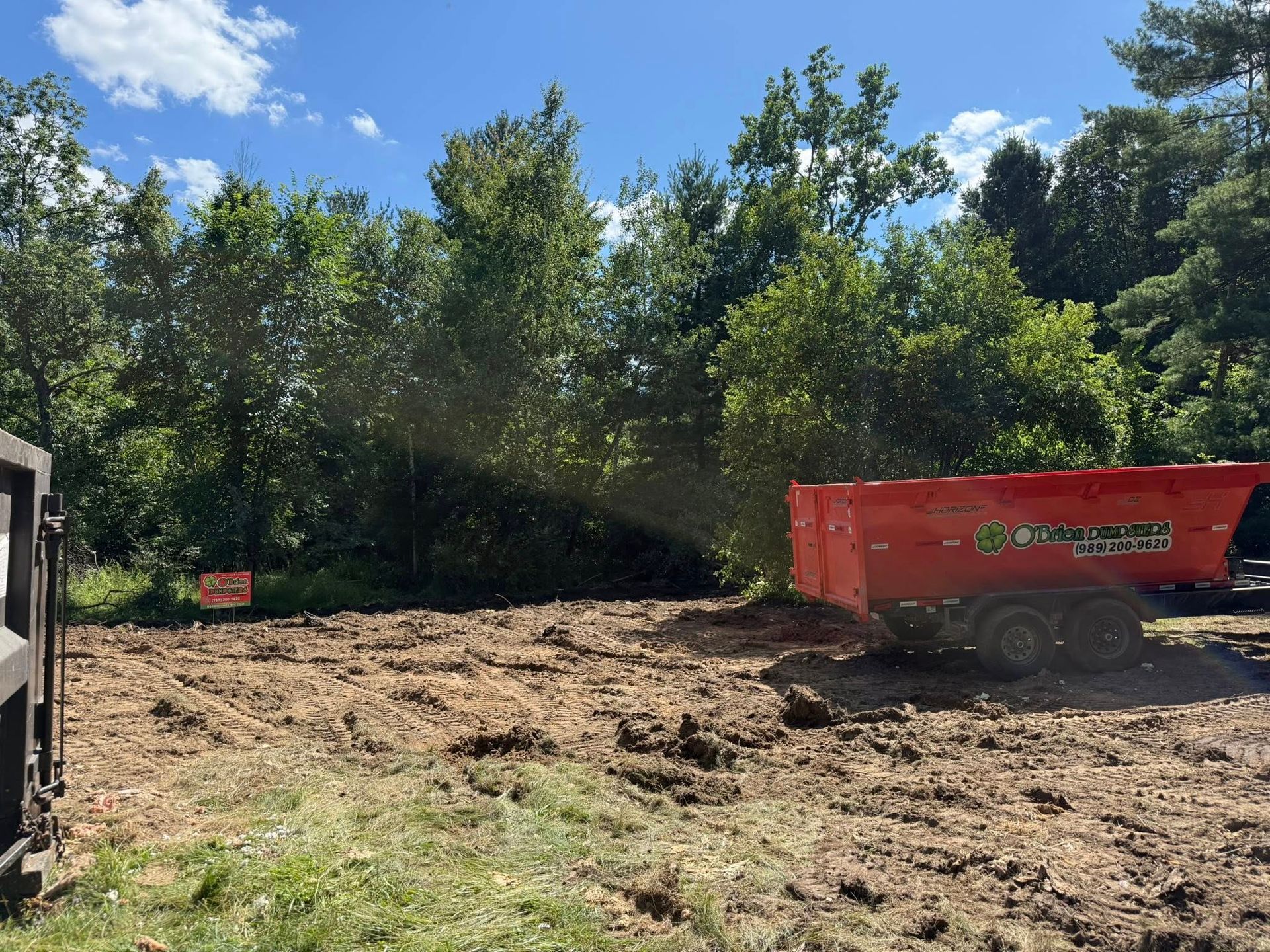 A cleared plot of land with an orange trailer, backed by trees under a blue sky.