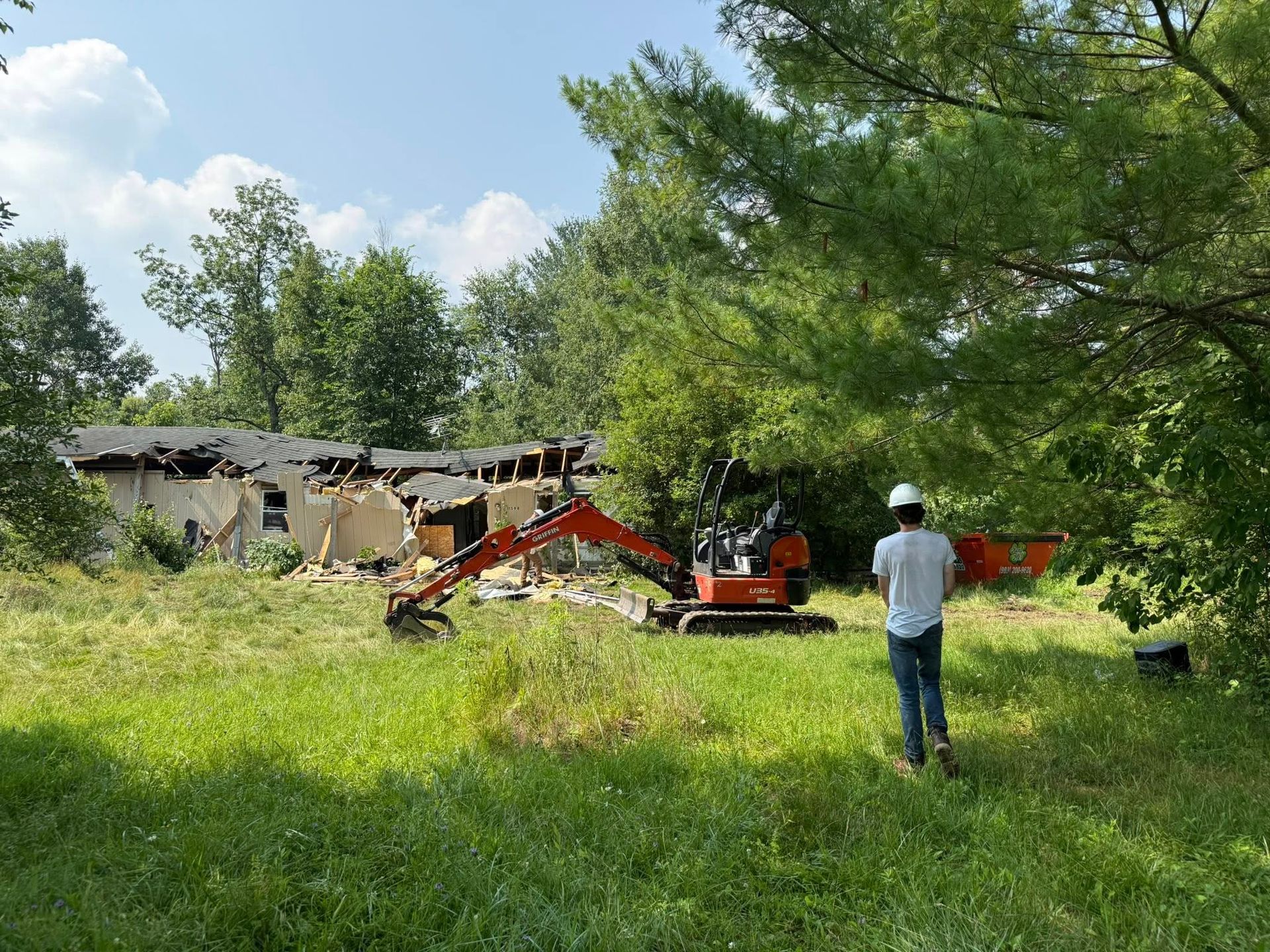 Excavator demolishing a building on a grassy lot. A person in a hard hat watches. Overcast sky and trees.
