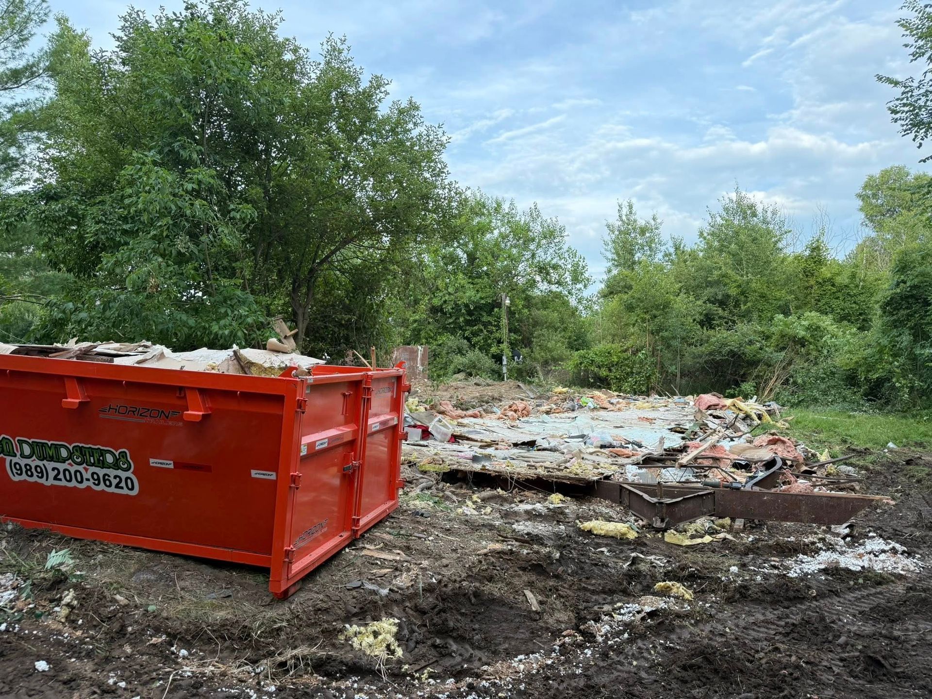 Red dumpster next to debris of a demolished building; trees in background; cloudy sky.