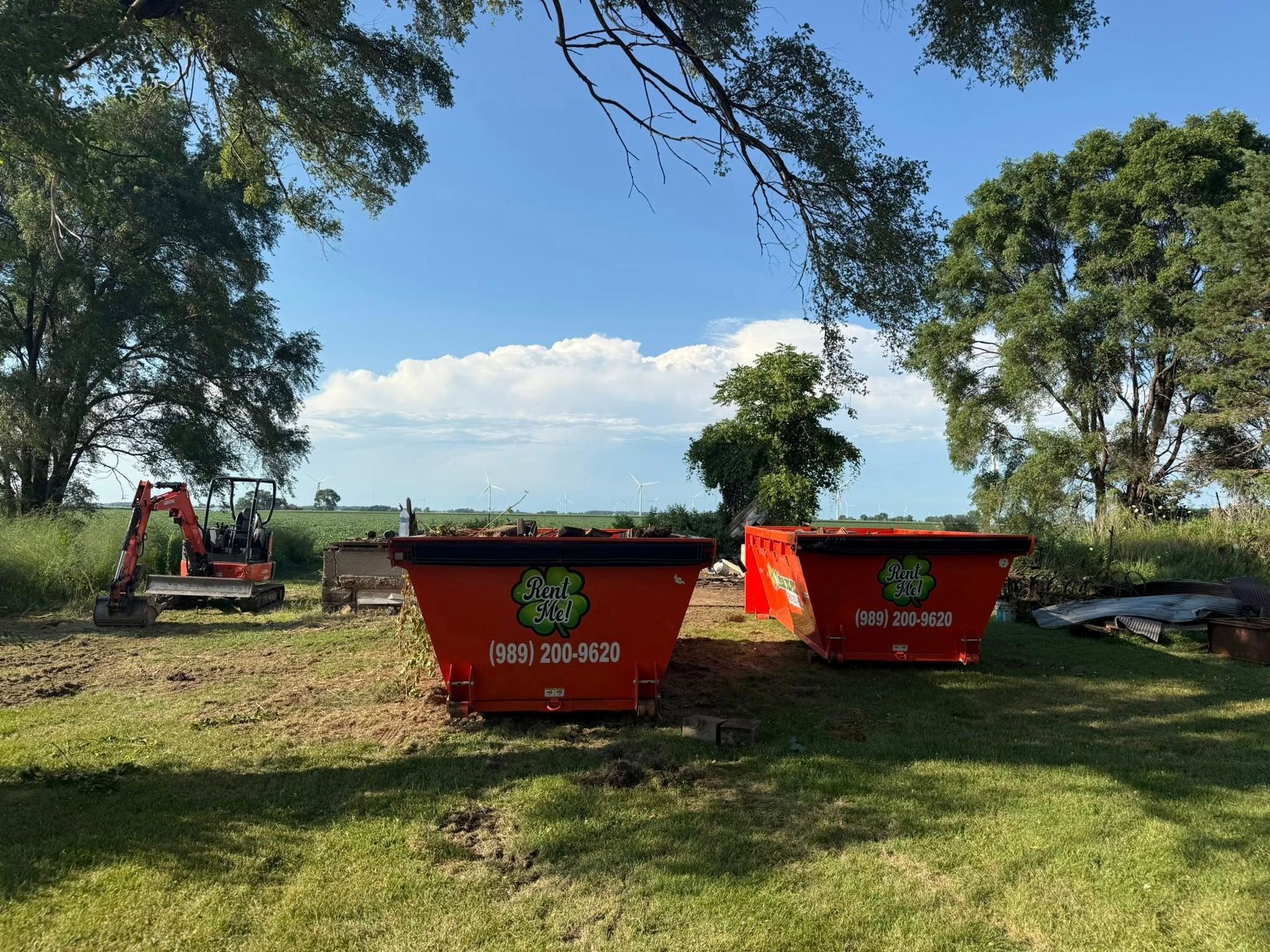Two orange dumpsters and small excavator on a grassy field under a blue sky.