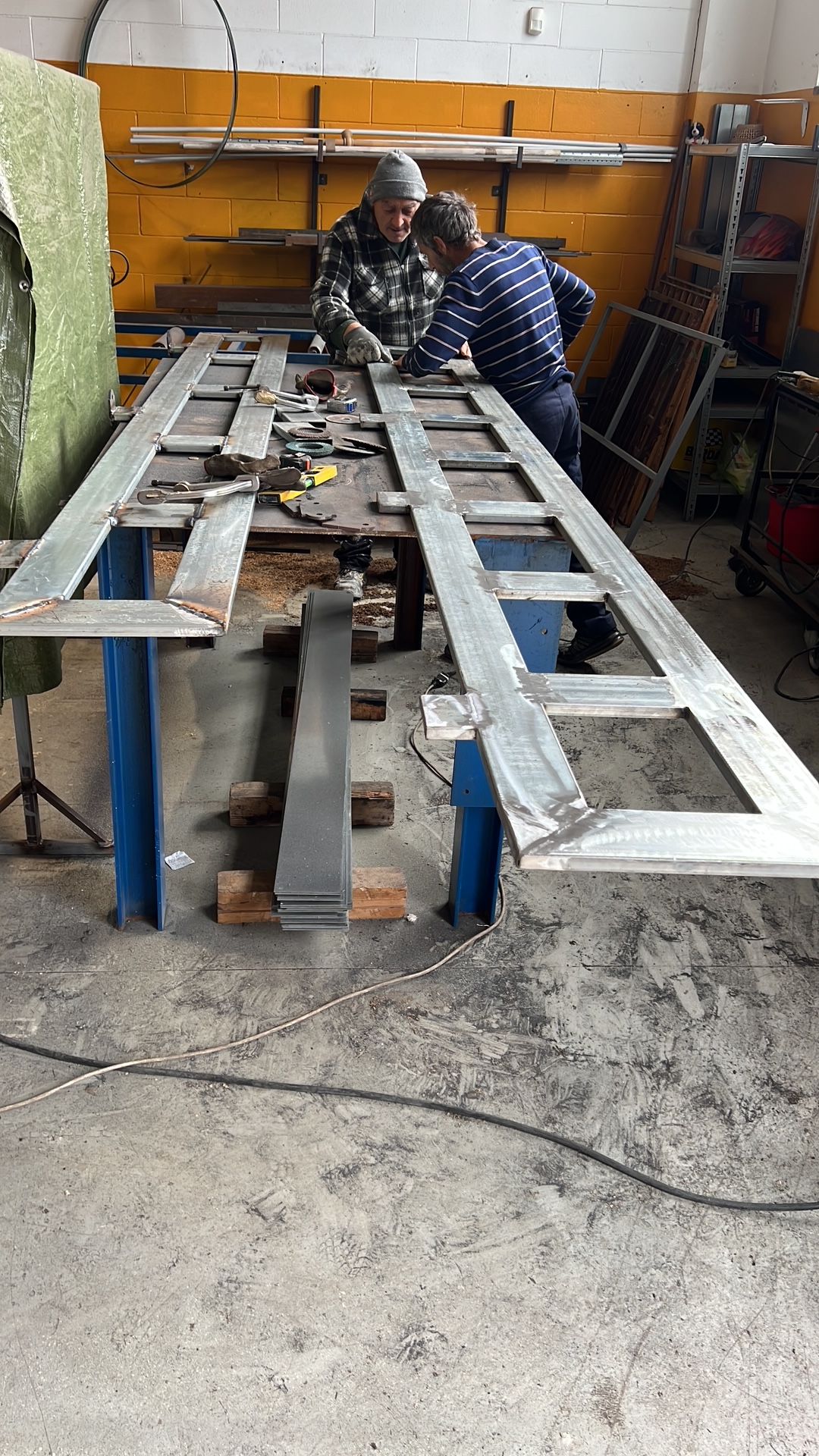 Due operai assemblano una lunga struttura di legno in un'officina, con scaffali e attrezzi alle loro spalle.