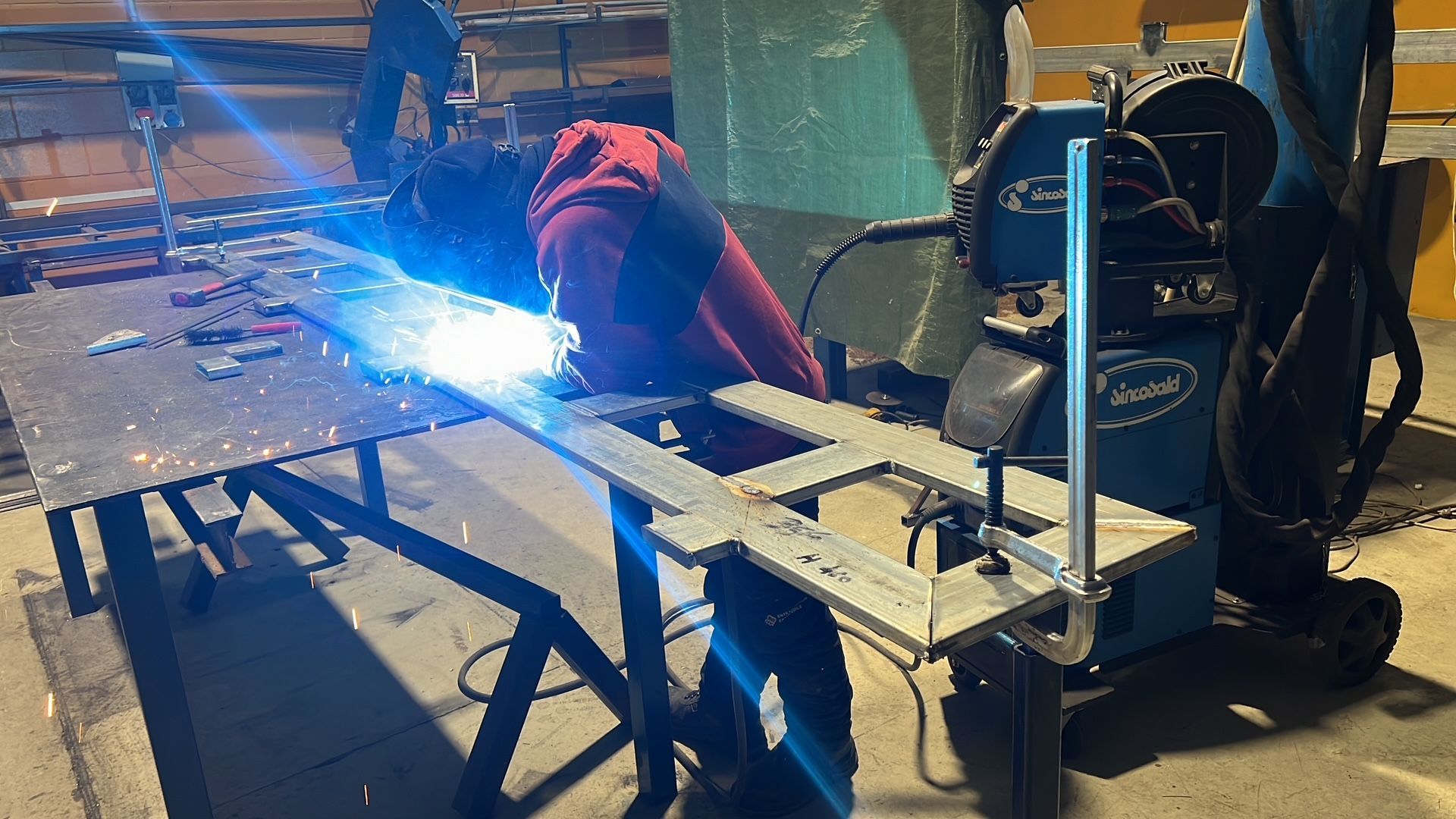 In un'officina, una persona salda pezzi di metallo su un banco da lavoro, producendo scintille di un blu brillante.