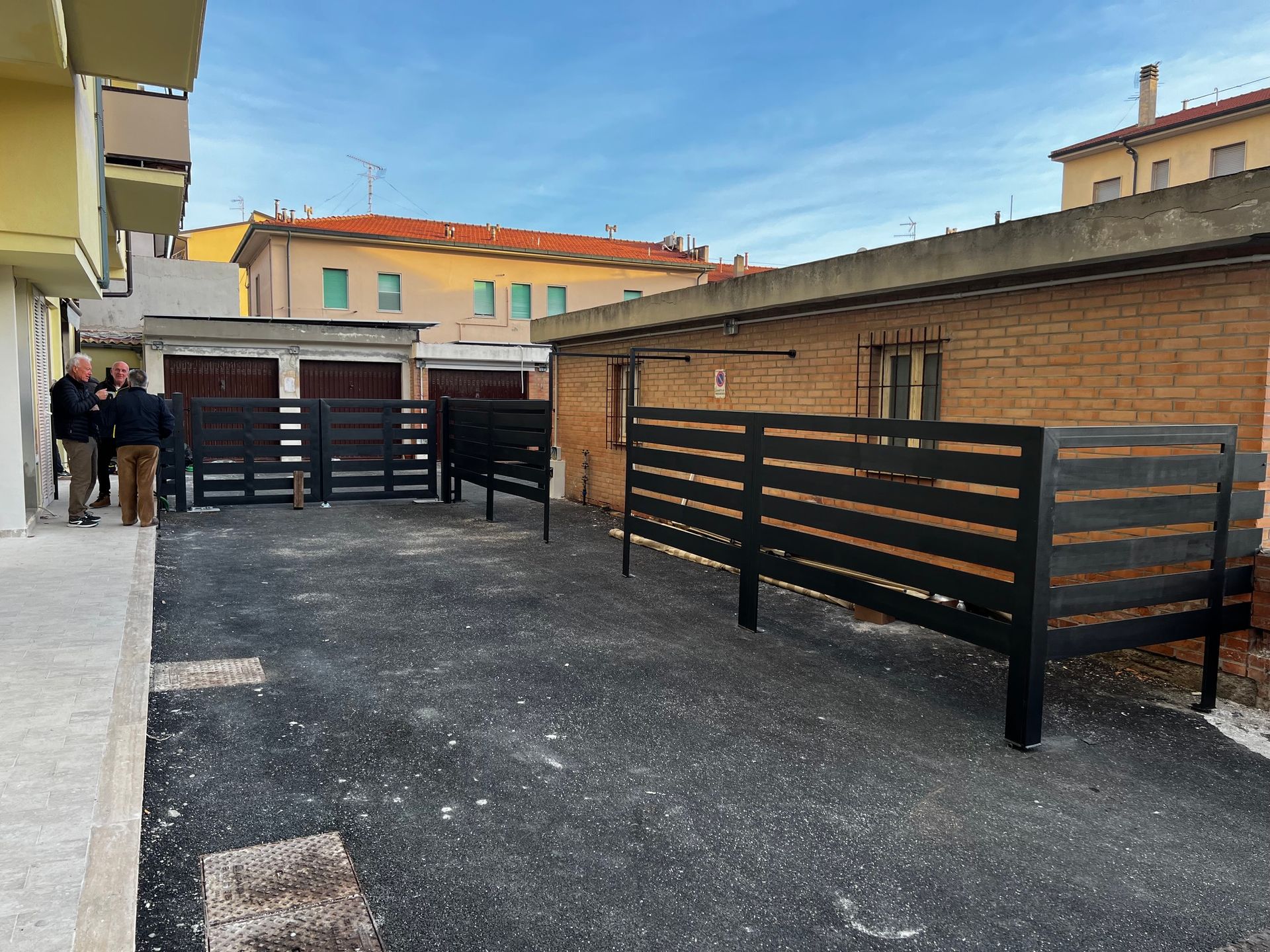 Cortile esterno con recinzione in metallo nero, edifici in mattoni e due persone vicino a un cancello.