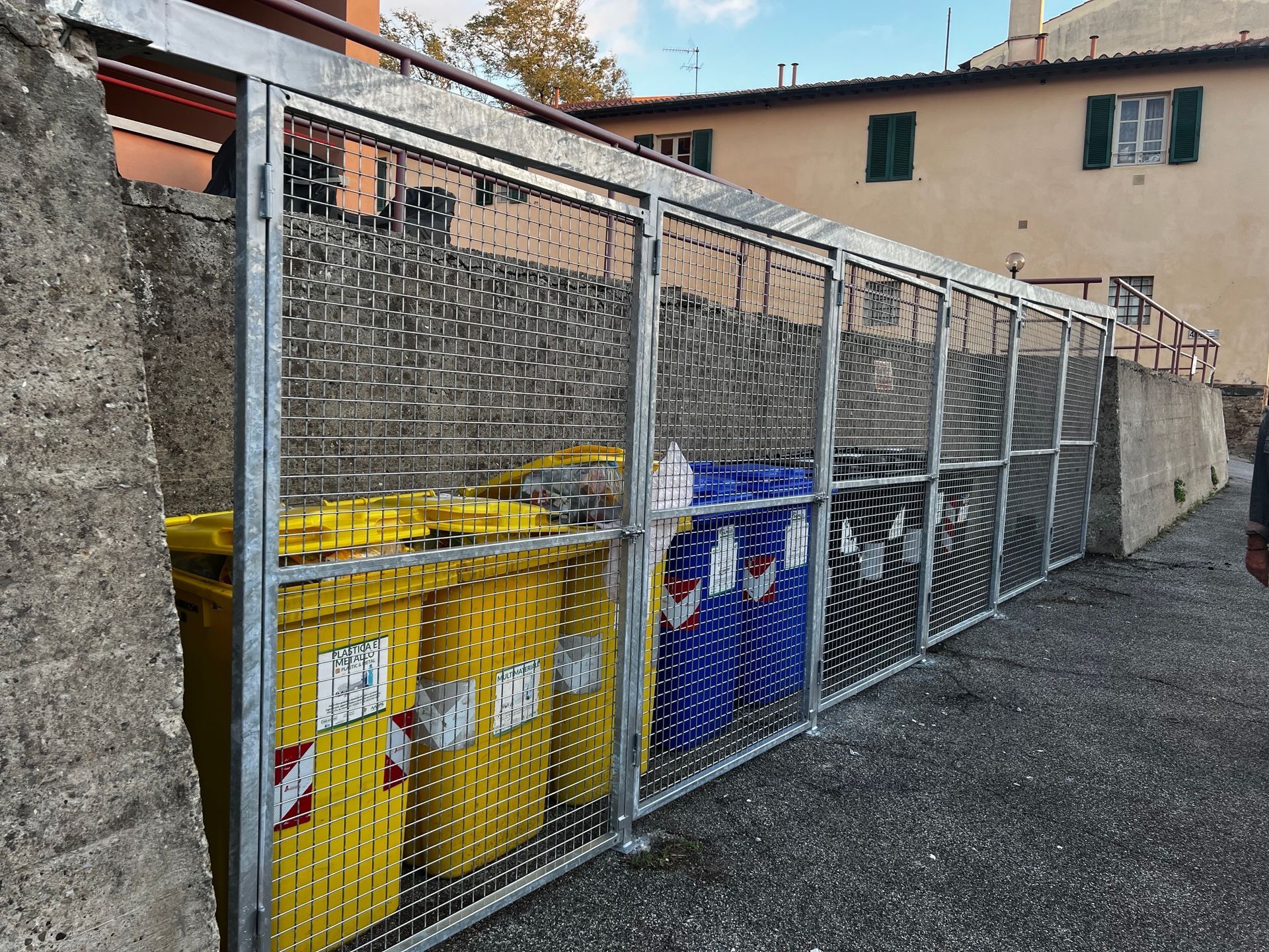 Una fila di contenitori per la raccolta differenziata, recintati e comprendenti contenitori gialli e blu, si trova accanto a un edificio e a un sentiero di ghiaia.