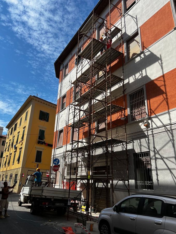 Impalcature su un palazzo residenziale rosso e bianco con operai, veicoli parcheggiati e un cielo azzurro.