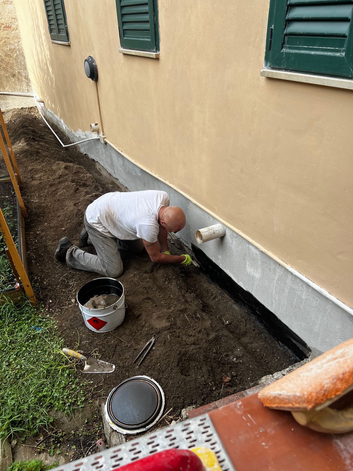 Una persona è inginocchiata accanto alle fondamenta di una casa, mentre lavora con un attrezzo in una trincea di terra vicino al muro.