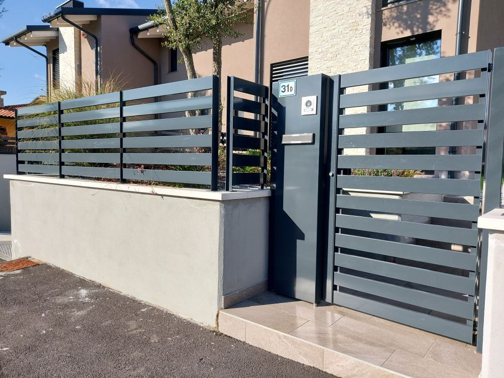 Ingresso di una casa moderna con recinzione a doghe grigie, cancello scuro e gradini piastrellati illuminati dalla luce del sole.
