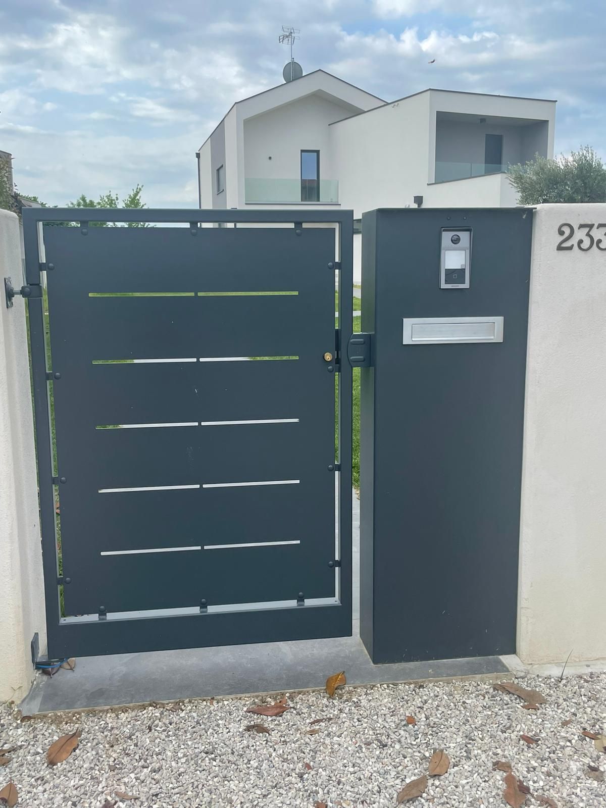 Ingresso di casa moderno con cancello grigio scuro, muri bianchi e vialetto di ghiaia.