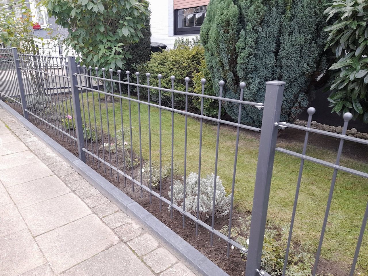 Recinzione metallica a lato di un marciapiede lastricato, che delimita un piccolo giardino erboso con arbusti e alberi ad alto fusto.