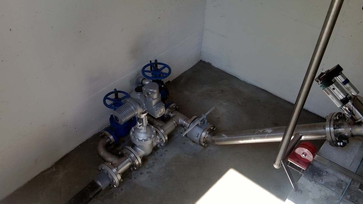 Valvole e tubature industriali in un angolo, con comandi a volantino blu su un pavimento di cemento.