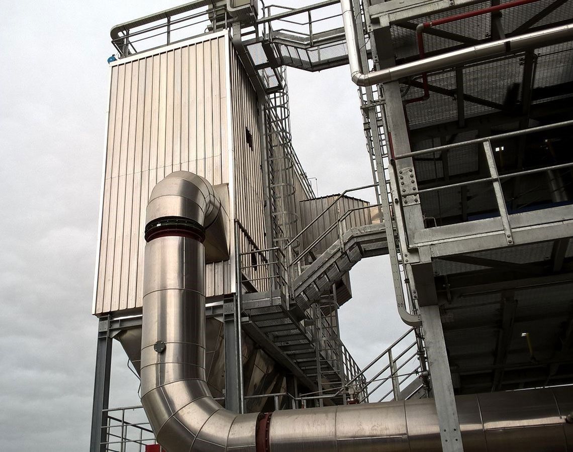 Stabilimento industriale con grandi condotti argentati, scale metalliche e un'alta torre beige che si staglia contro un cielo grigio.