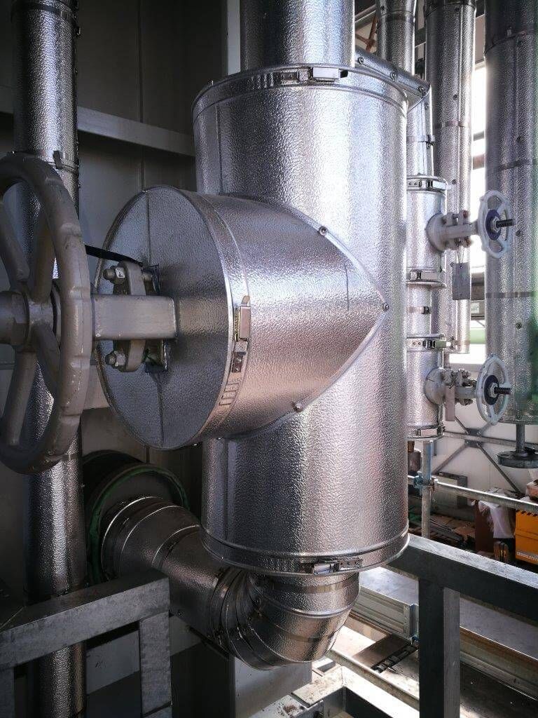 Condotti e tubazioni metalliche industriali con un grande serbatoio cilindrico argentato in un ambiente di fabbrica.