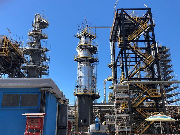 Torri e tubature di una raffineria industriale si stagliano contro un cielo azzurro limpido, con un edificio blu in primo piano.