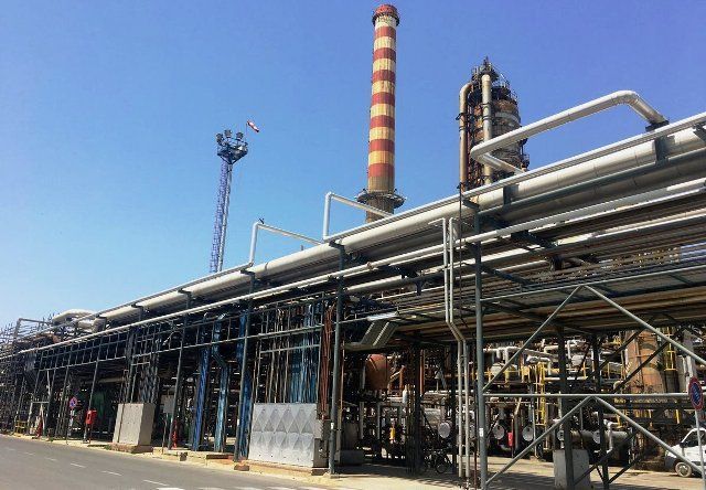 Impianto industriale con grandi tubi metallici, impalcature e un'alta ciminiera a strisce sotto un cielo azzurro limpido.