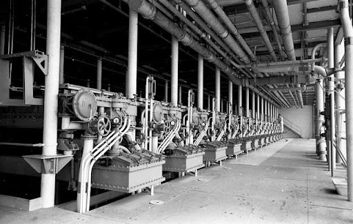 Interno di uno stabilimento industriale con una lunga fila di macchinari, tubature e attrezzature sotto un tetto metallico.
