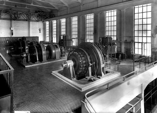 Foto in bianco e nero dell'interno di una centrale elettrica storica, con grandi turbine e alte finestre.