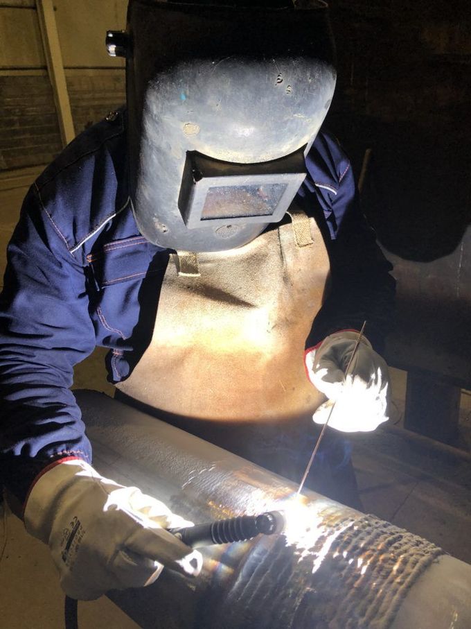 Un saldatore con indumenti protettivi sta saldando un tubo di metallo, con scintille che volano in un'officina poco illuminata.