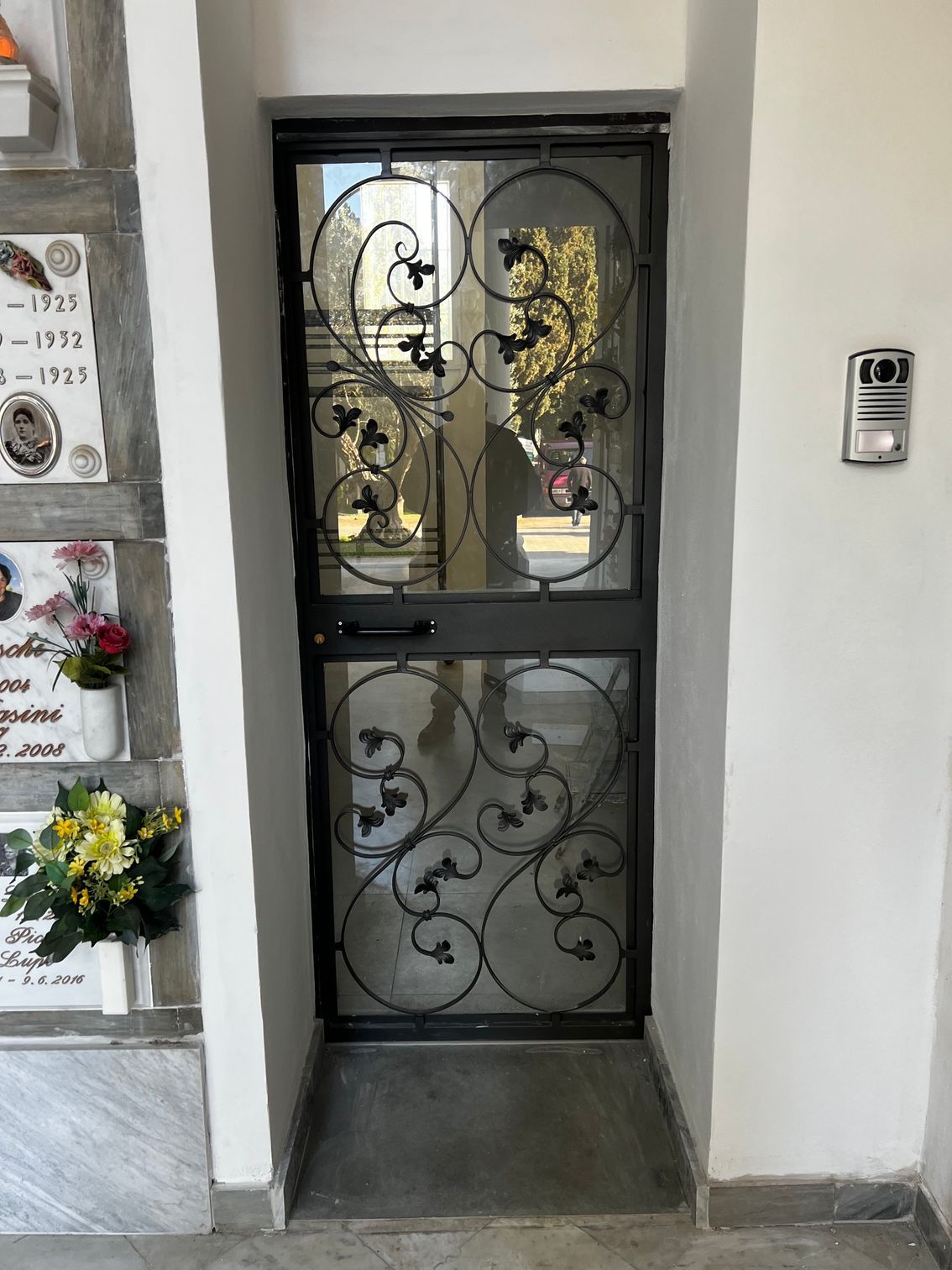 Cancello in ferro battuto nero all'ingresso di un edificio, con citofono a muro sulla destra.