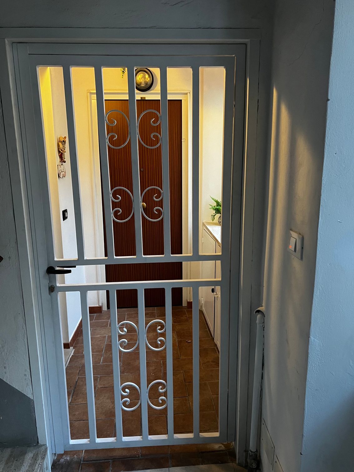 Cancello di sicurezza bianco in uno stretto ingresso, oltre il quale si intravede una porta d'ingresso in legno.