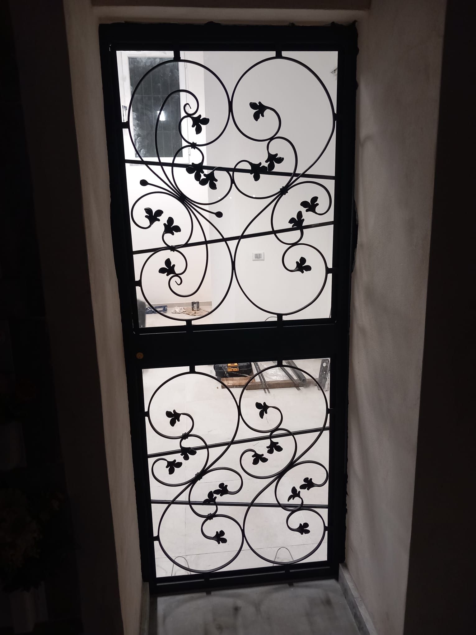 Griglia decorativa in ferro battuto nero su una parete bianca, con motivi floreali a spirale.
