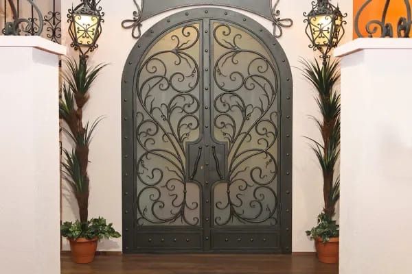 Doppie porte in ferro battuto decorate, fiancheggiate da lanterne e piante in vaso, su un ingresso piastrellato.