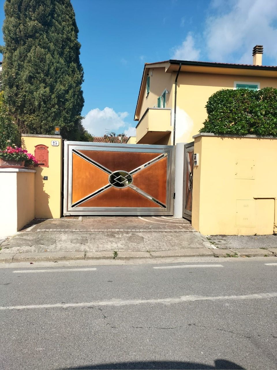 Cancello carrabile residenziale con muri gialli e una casa alle sue spalle, su una strada soleggiata.