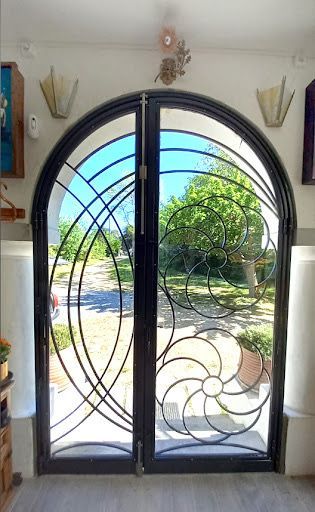 Doppia porta d'ingresso in ferro battuto con pannelli in vetro ad arco, che si apre su un sentiero soleggiato in giardino.