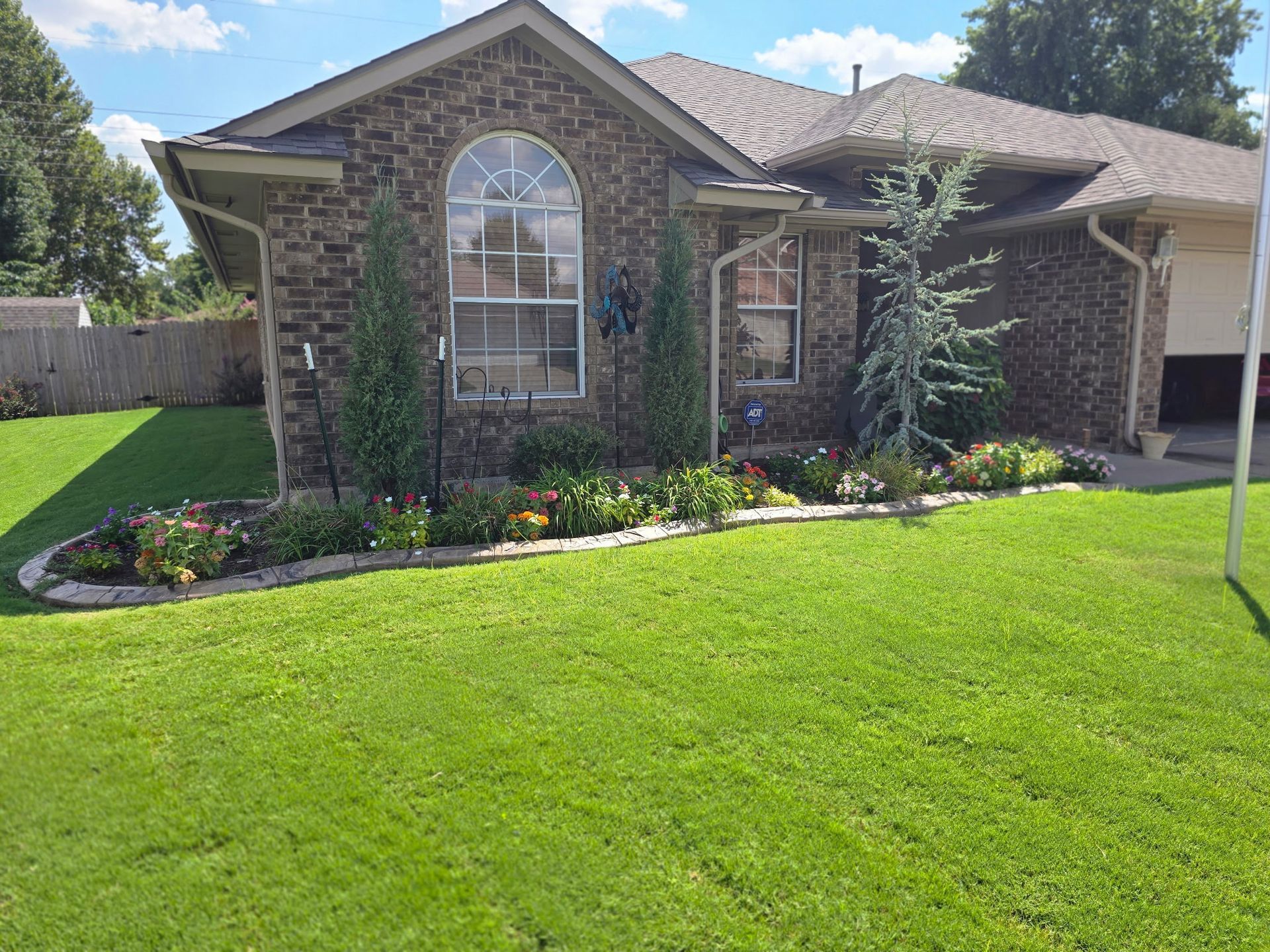 A brick house with a landscaped garden bed featuring small evergreens and flowers in front of a green lawn.