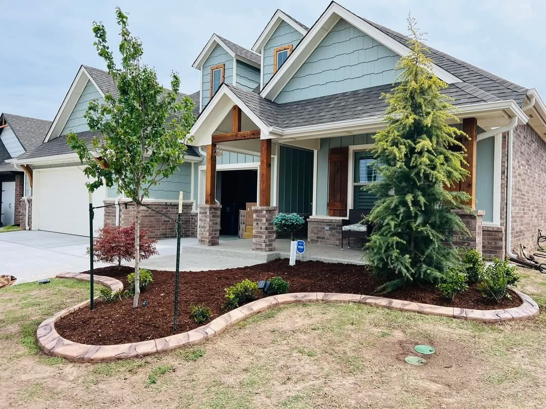 A light blue house with stone accents and a front porch, featuring a landscaped garden bed with mulch and young trees.