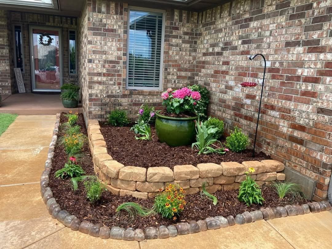 A terraced garden bed with a large green pot, bright pink flowers, and decorative stones in front of a brick house.