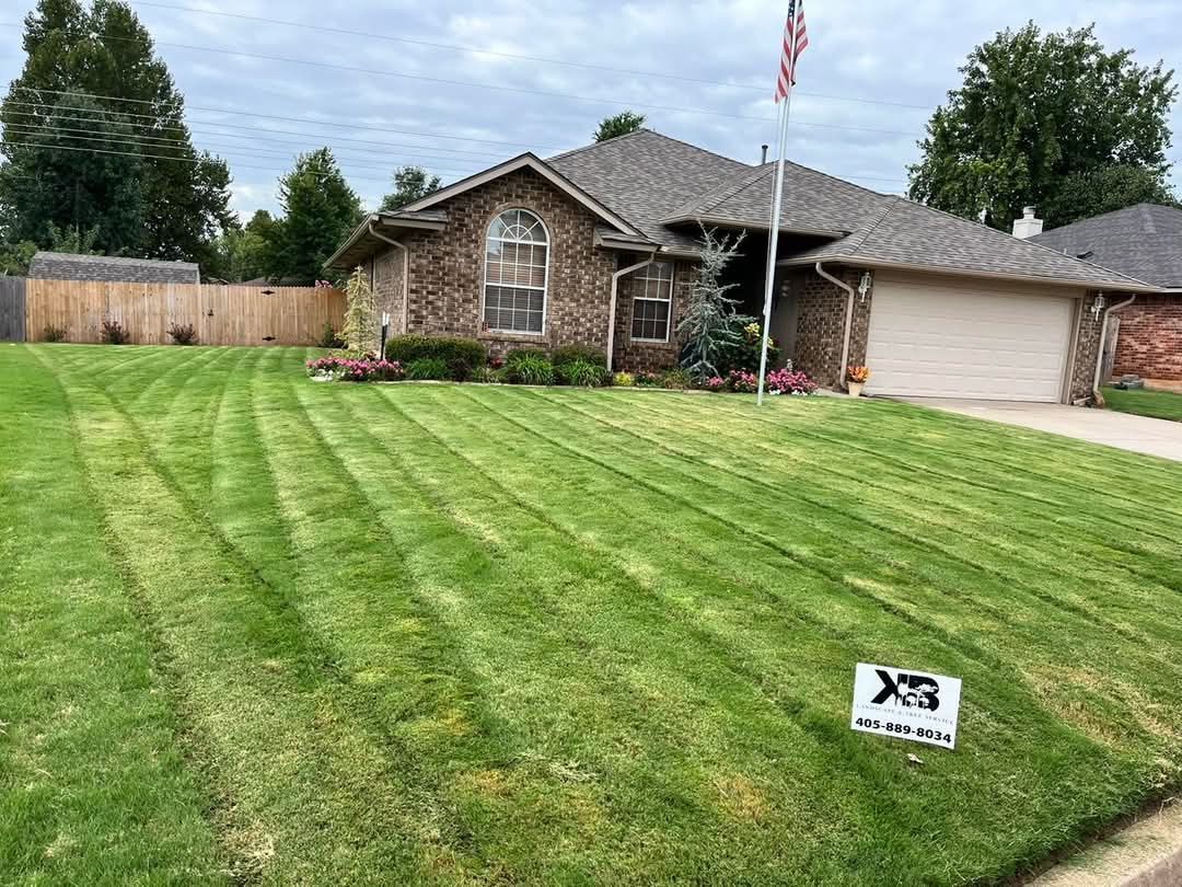 A single-story brick house with a neatly mowed lawn, a paved driveway, and an American flag in the front yard.