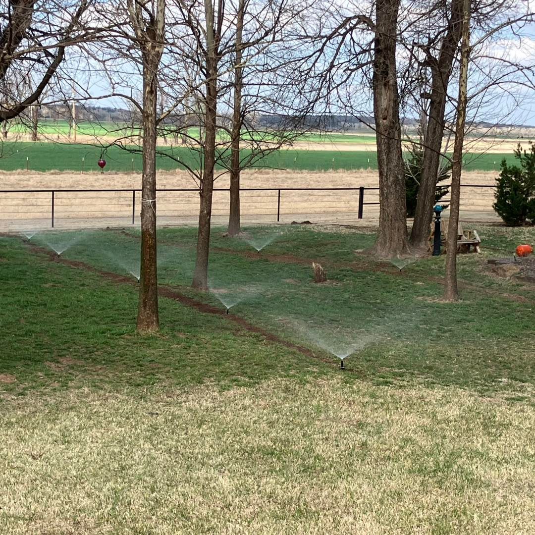 Multiple sprinklers spray a misty water arc over a green lawn in front of bare trees and a distant rural field.