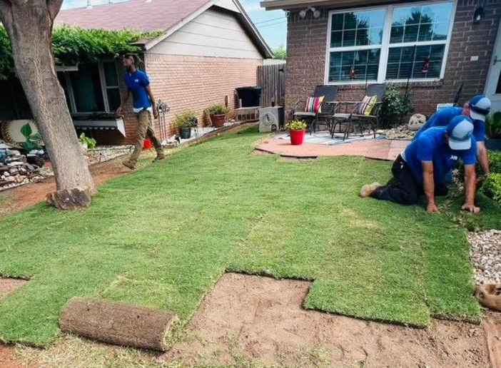 Workers install rolls of fresh green sod on a residential lawn, with one person kneeling to position a piece.