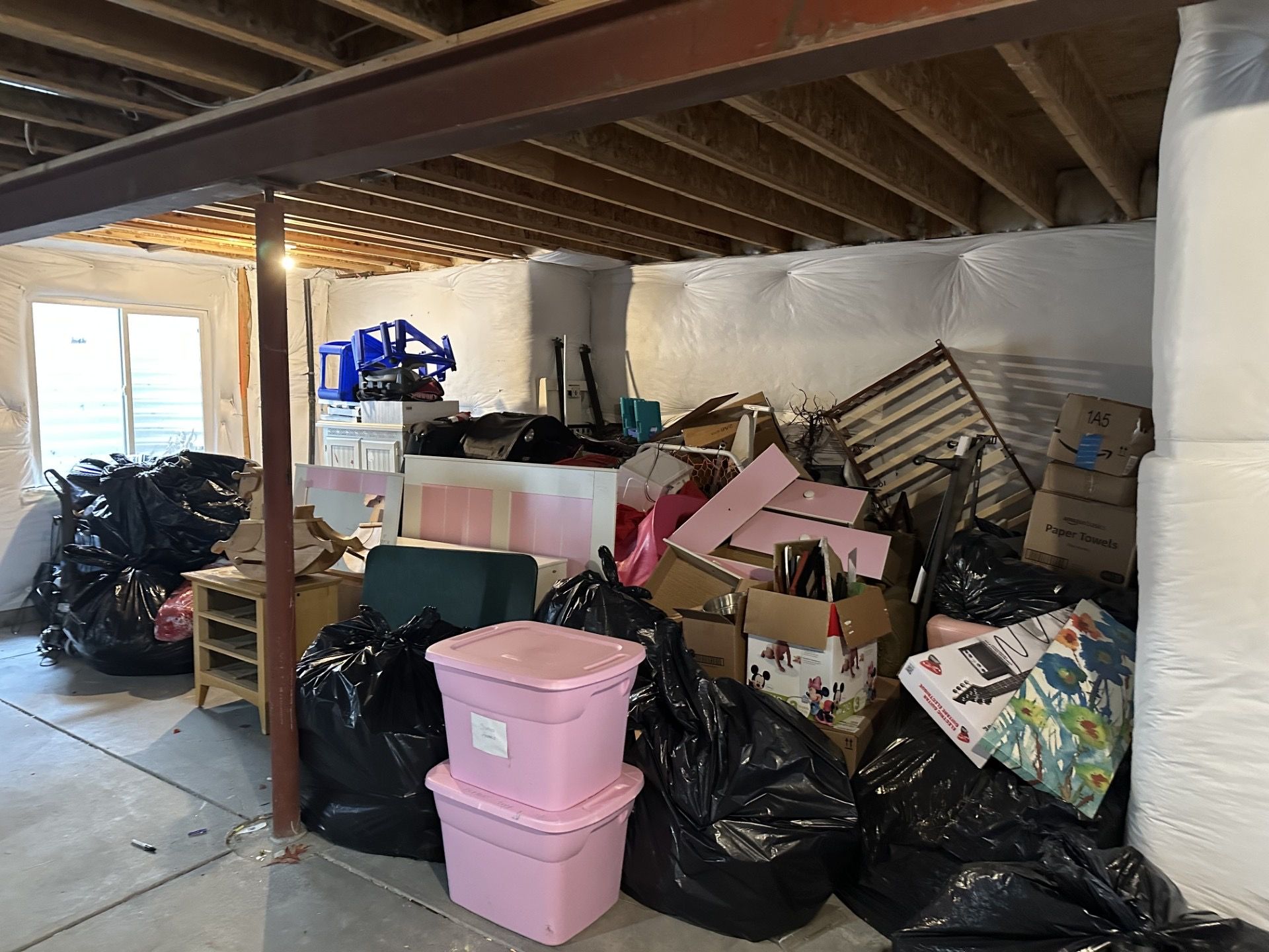 Cluttered basement: Black trash bags, pink bins, boxes, and debris piled against the wall.