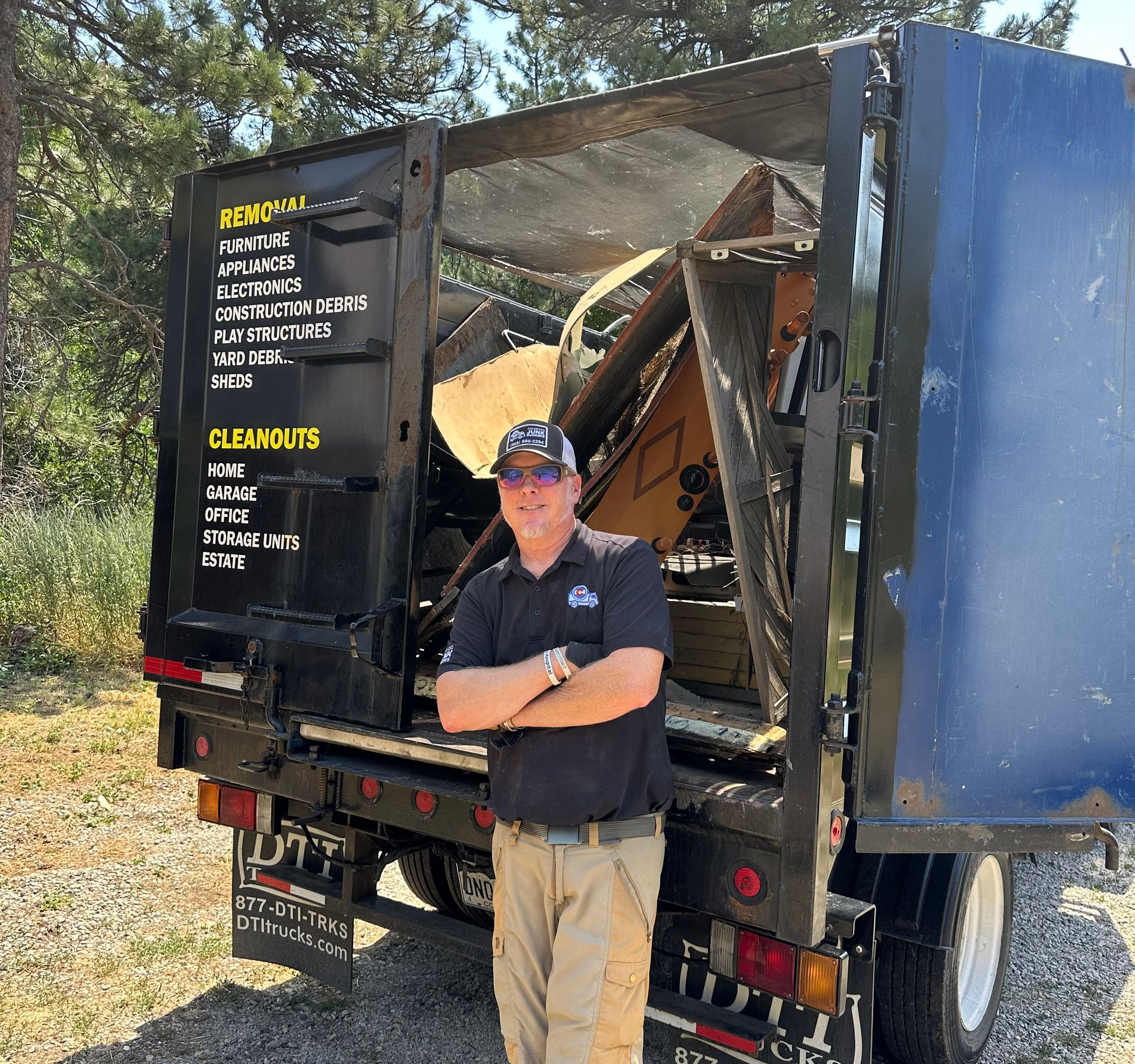 Owner Tom stands with arms crossed in front of a his junk truck filled with junk, displaying a list of services.