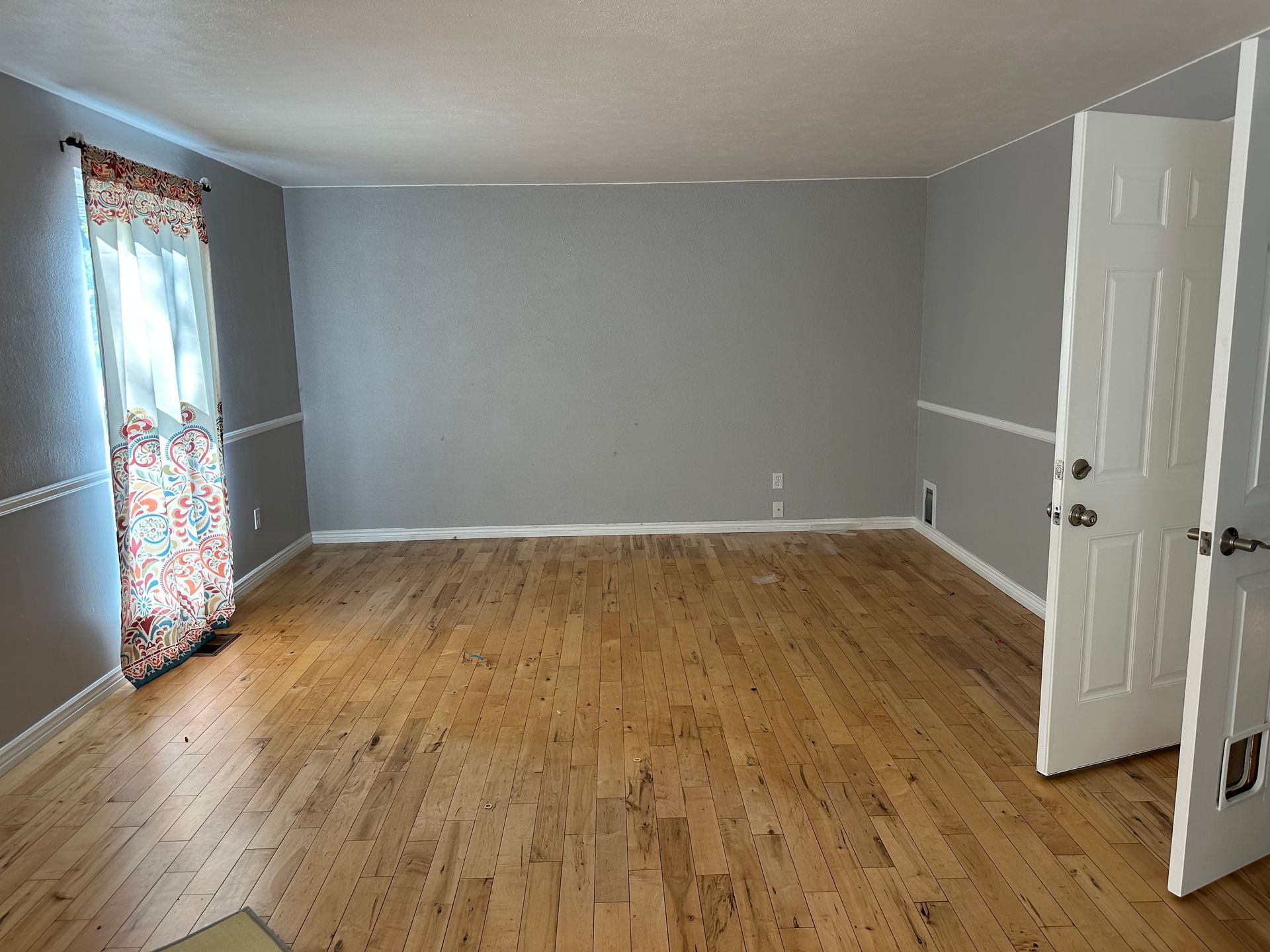 Empty room with hardwood floors, gray walls, white trim, and a partially open door. Sunlight streams through a window.