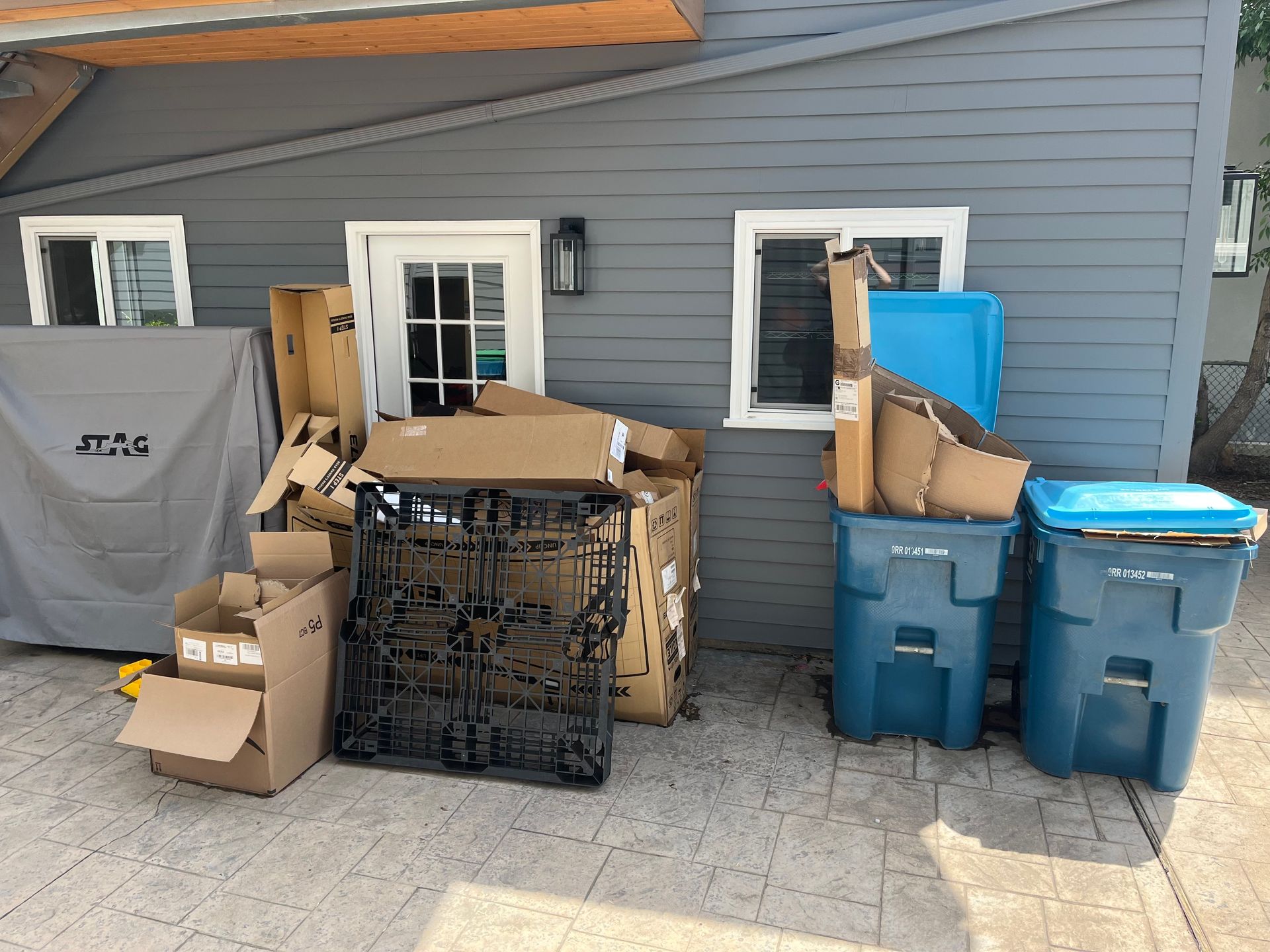 Cardboard boxes, trash bins, and a grill cover next to a gray building. A before picture taken in Boulder, CO.