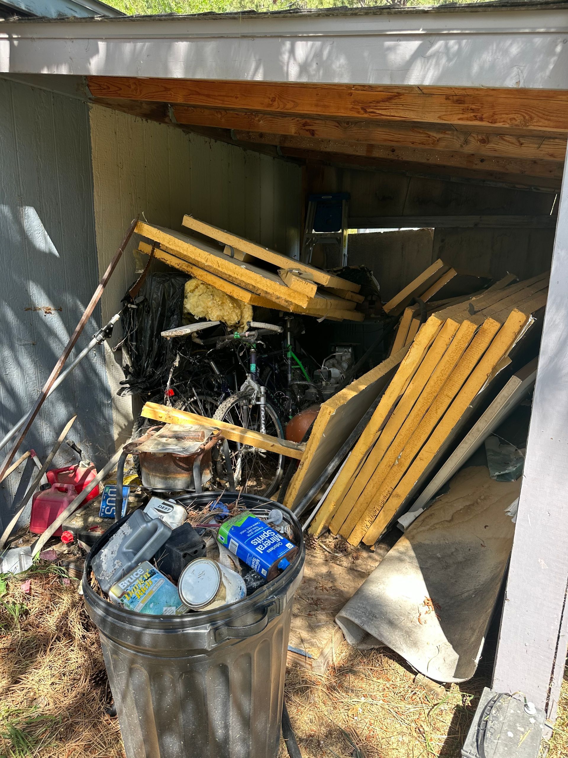 Overflowing trash can and discarded items clutter a shed containing bicycles and wood scraps.