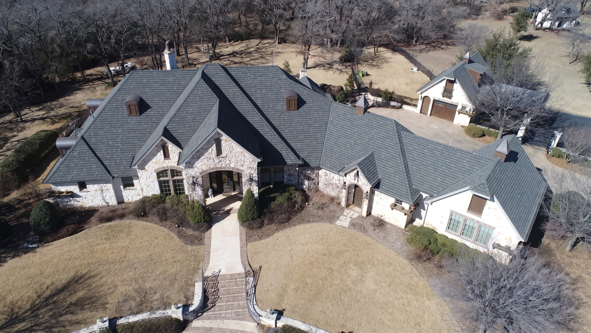 Aerial view of a large stone house with a dark roof and landscaped front yard.