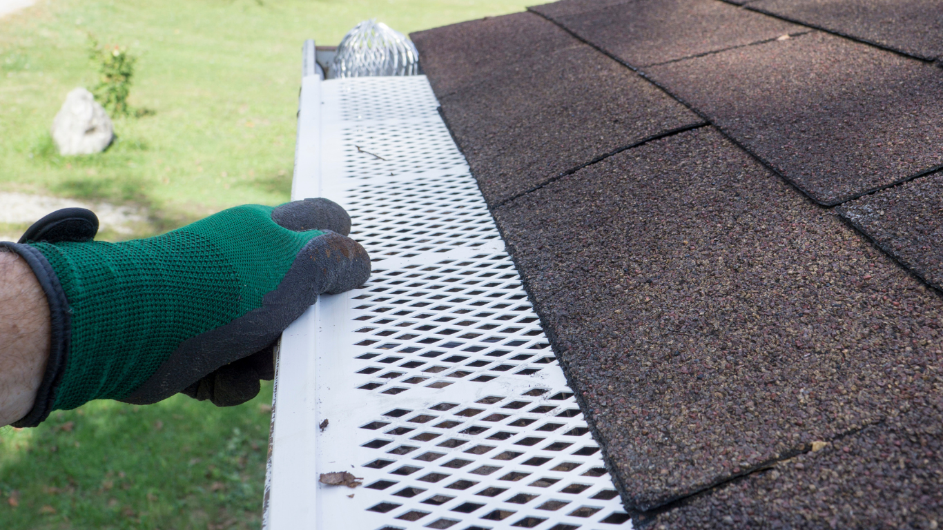 Gloved hand holding white roof gutter guard beside brown shingle roof edge