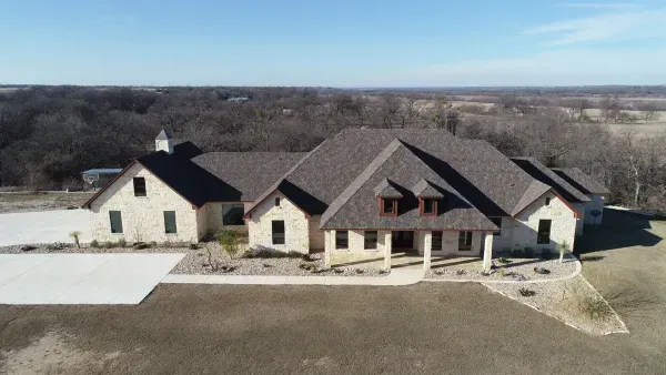 Aerial view of a large suburban house with a stone facade, dark roof, and wide front lawn.