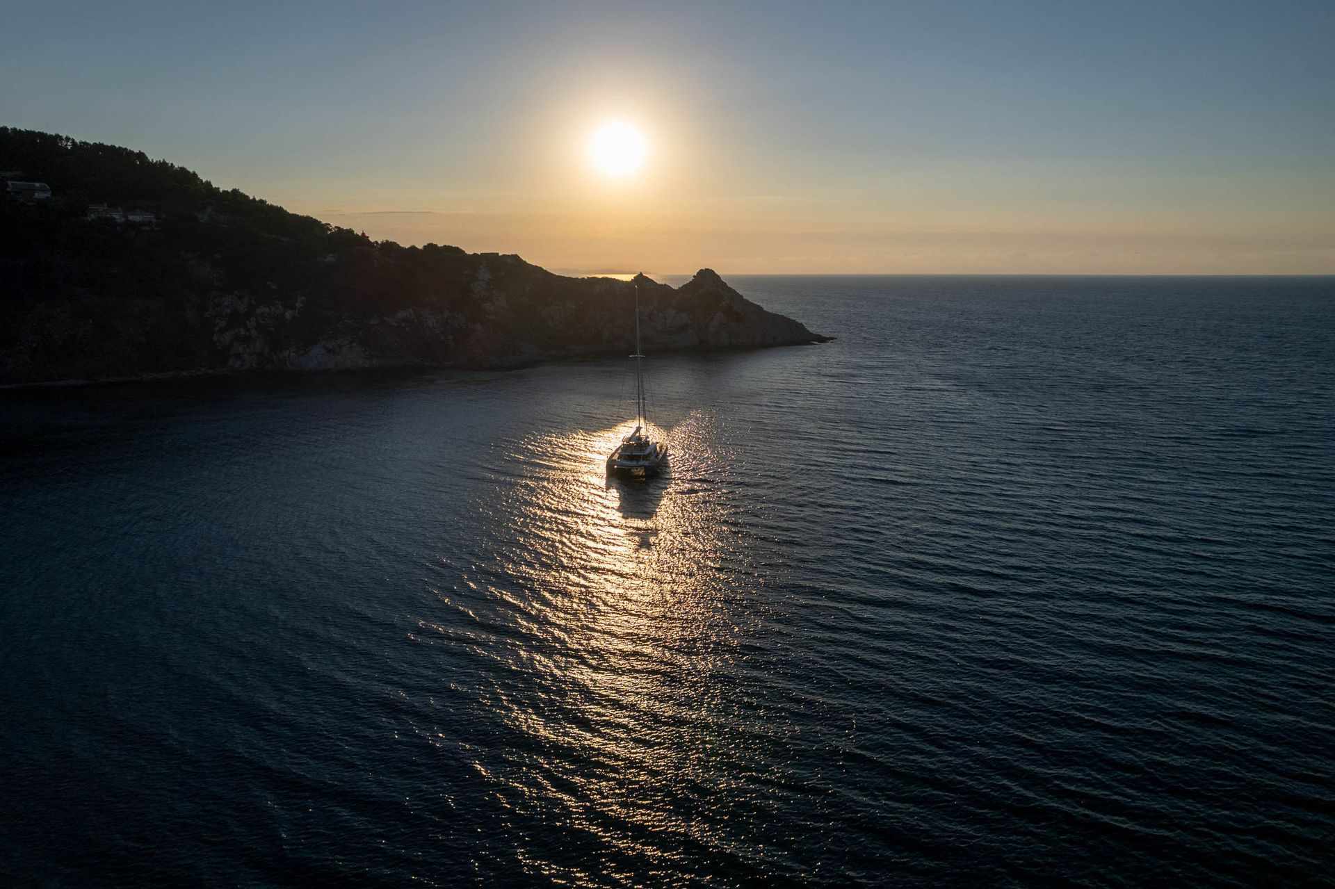 Distant catamaran in a calm sunset reflection between Mediterranean headlands
